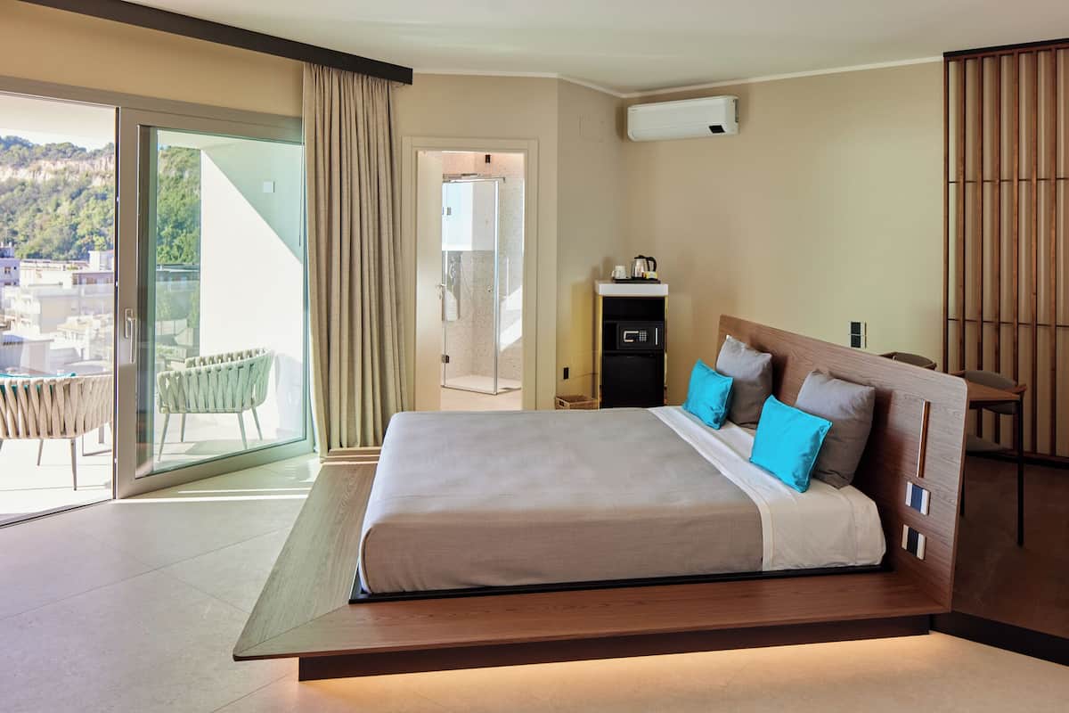 A modern hotel room with a large bed, a small desk, a chair, and a balcony with a view of the city.