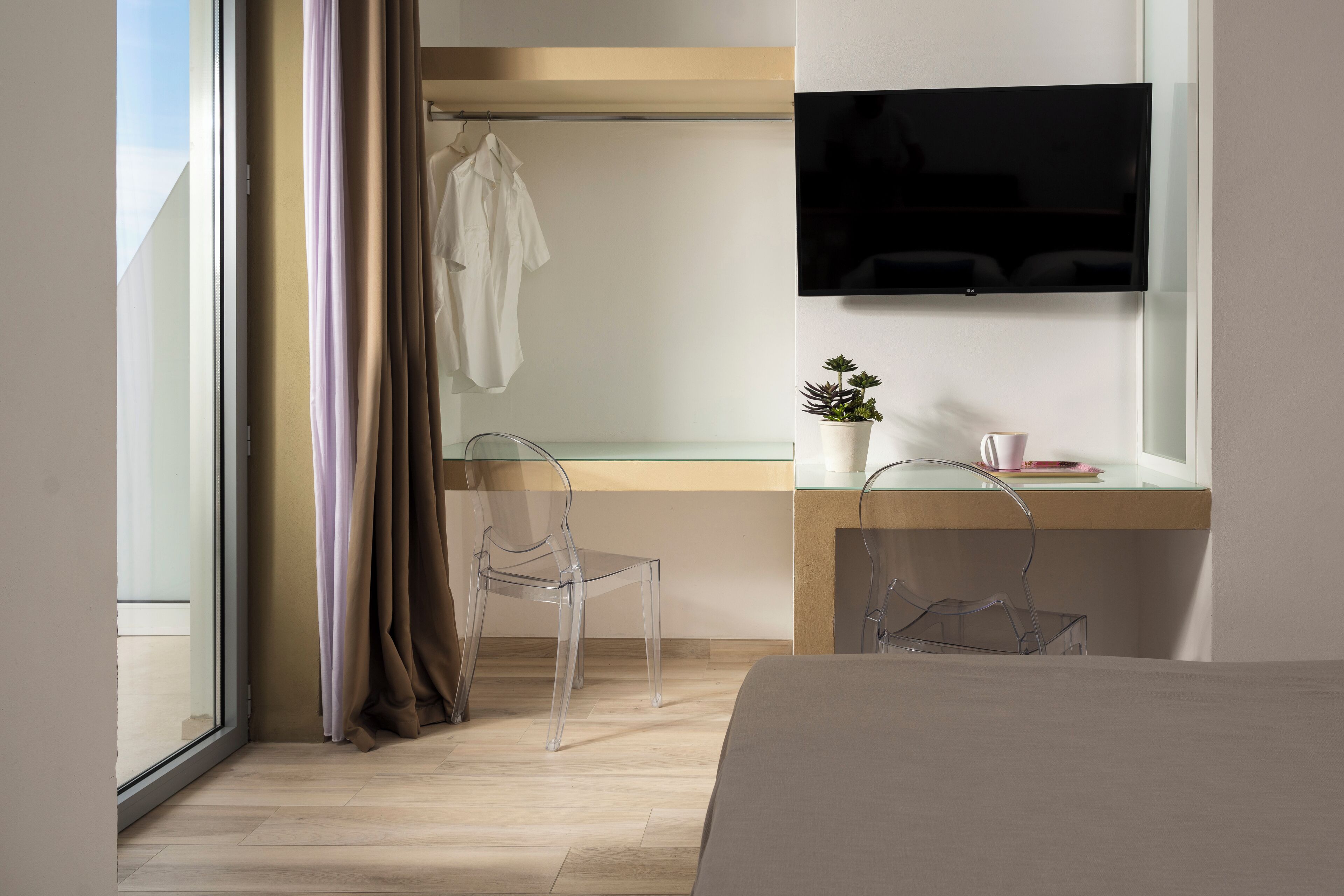 A modern hotel room with a bed, a desk with a TV, a chair, a potted plant, and a window with curtains.