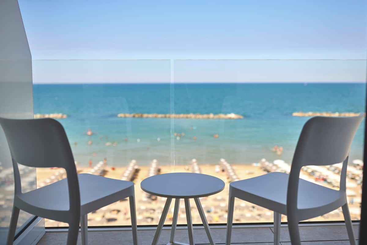 A balcony with two white chairs and a small table, overlooking a beach with umbrellas and a pier.