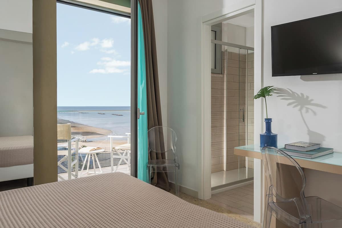 A bedroom with a large window overlooking a beach, a bed, a desk with a plant, and a TV on the wall.
