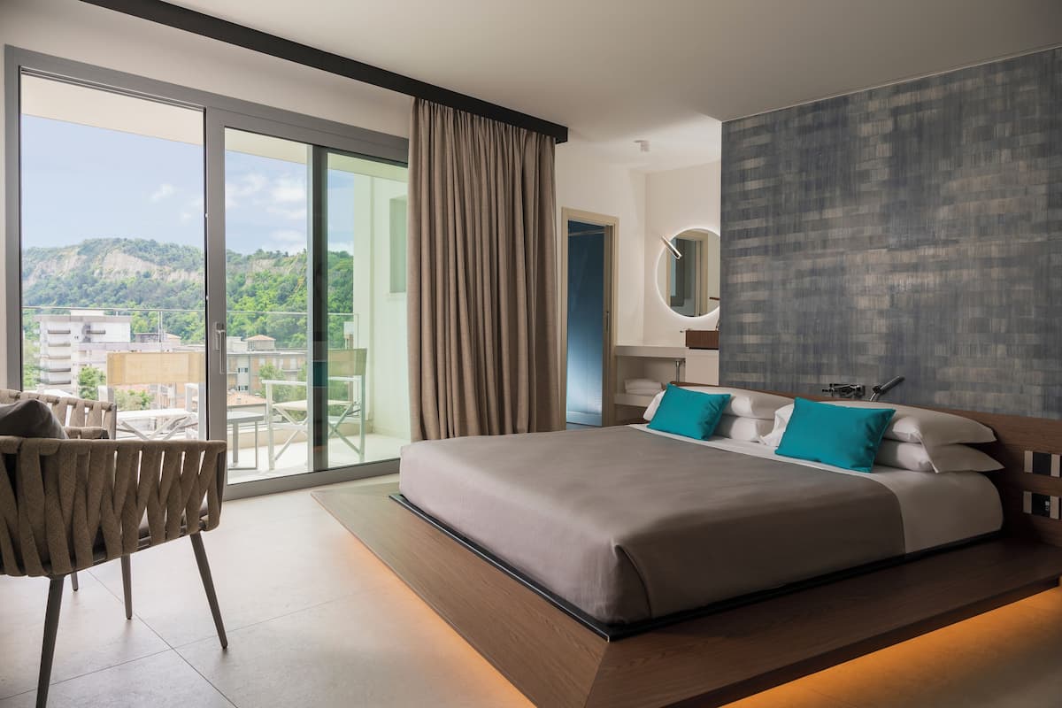 A modern hotel room with a large bed, a sitting area with a chair, a bathroom with a mirror, and a balcony with a view of the city.