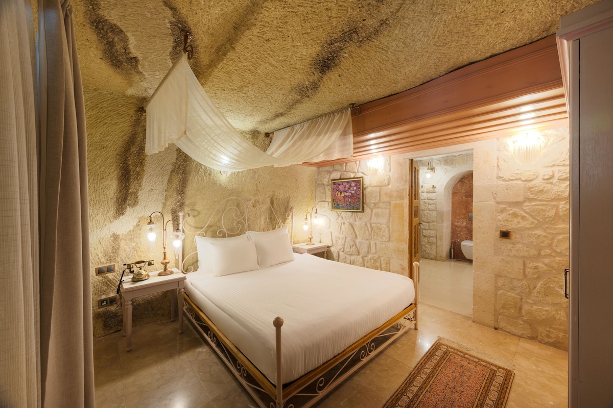 A bedroom with a stone wall, a bed with a canopy, a nightstand with a lamp, and a bathroom visible through an archway.