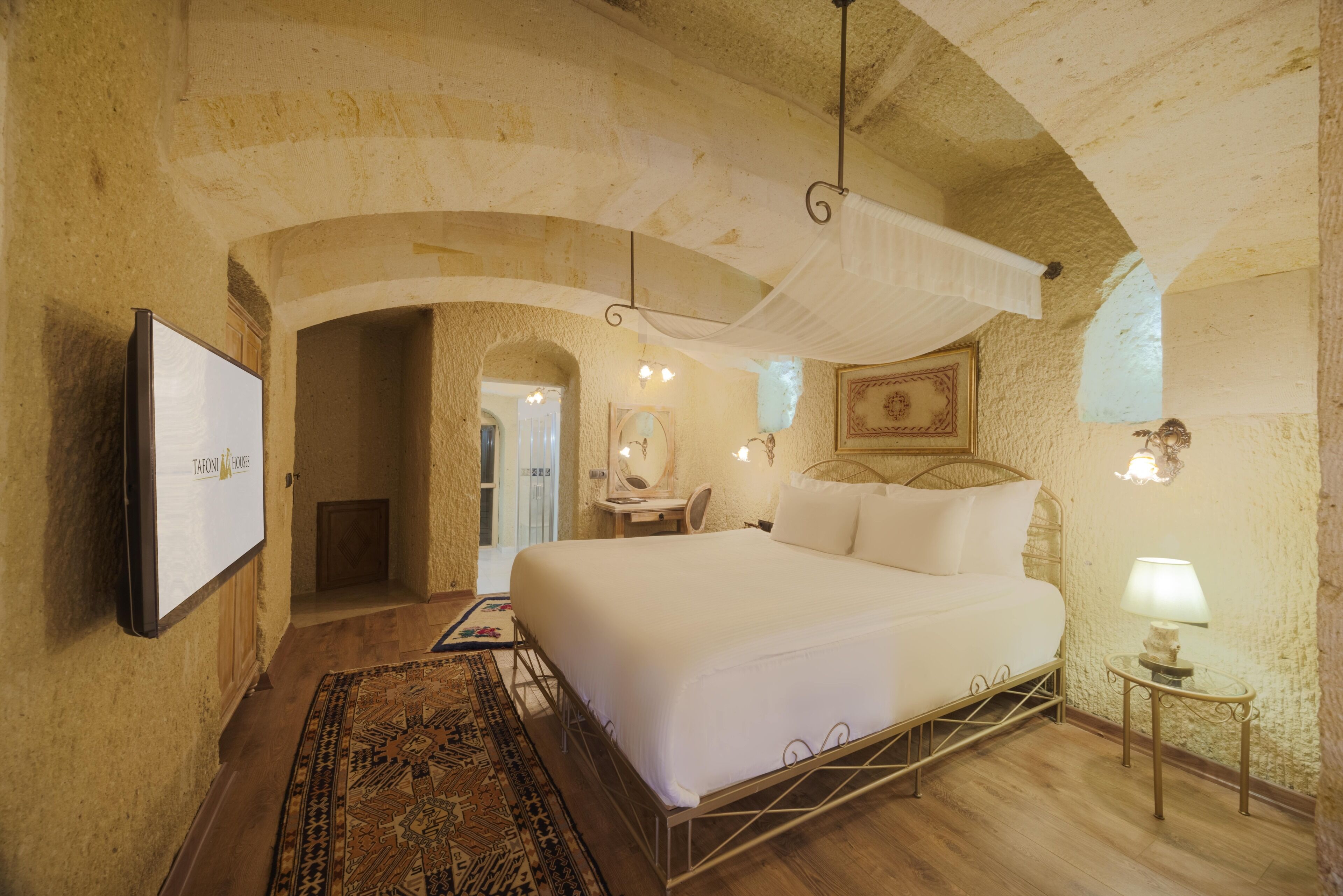 A bedroom with a large bed, a television on the wall, a small table with a lamp, and a patterned rug.