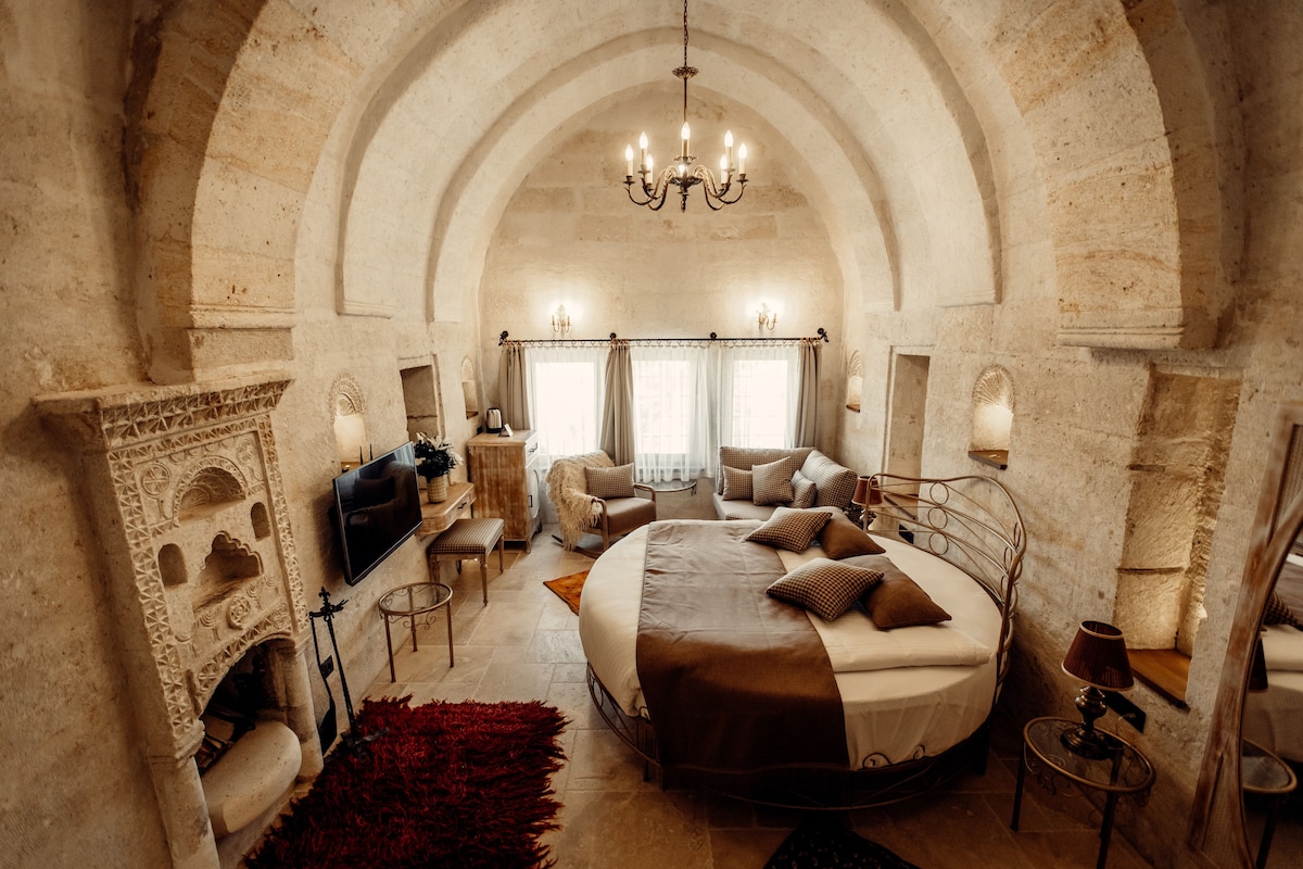 A bedroom with a stone arch ceiling, a chandelier, a bed with pillows, a television, and a fireplace.