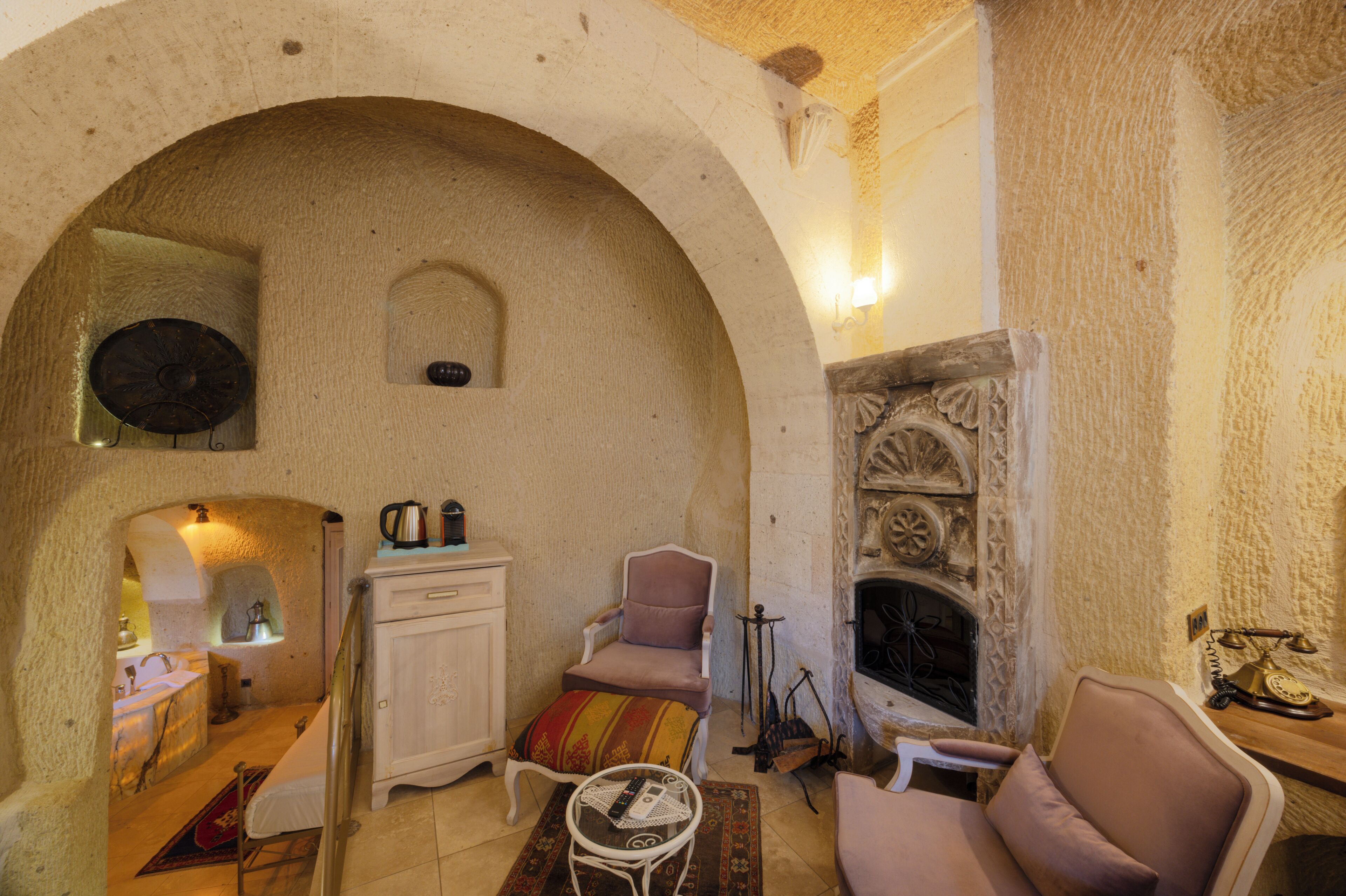 Un interior acogedor con un arco de piedra, una chimenea, un gabinete blanco, un sillón mullido y una pequeña mesa con un control remoto.