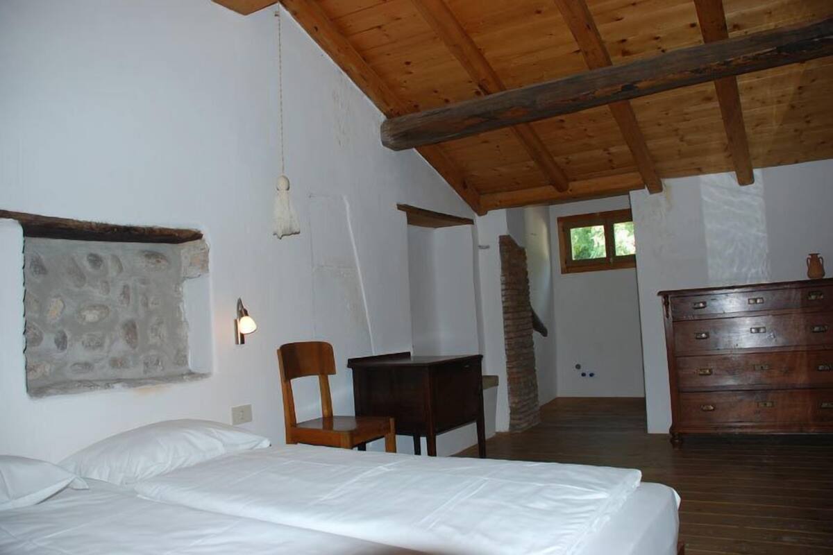 A room with a wooden ceiling, a stone wall, a bed, a chair, a dresser, and a small table.