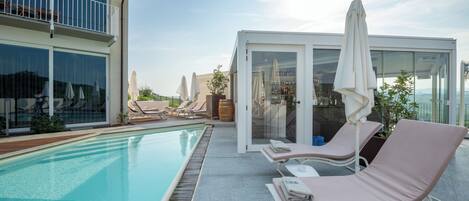 Zona de piscina con sillas de descanso, bar con paredes de cristal y vista a la piscina.