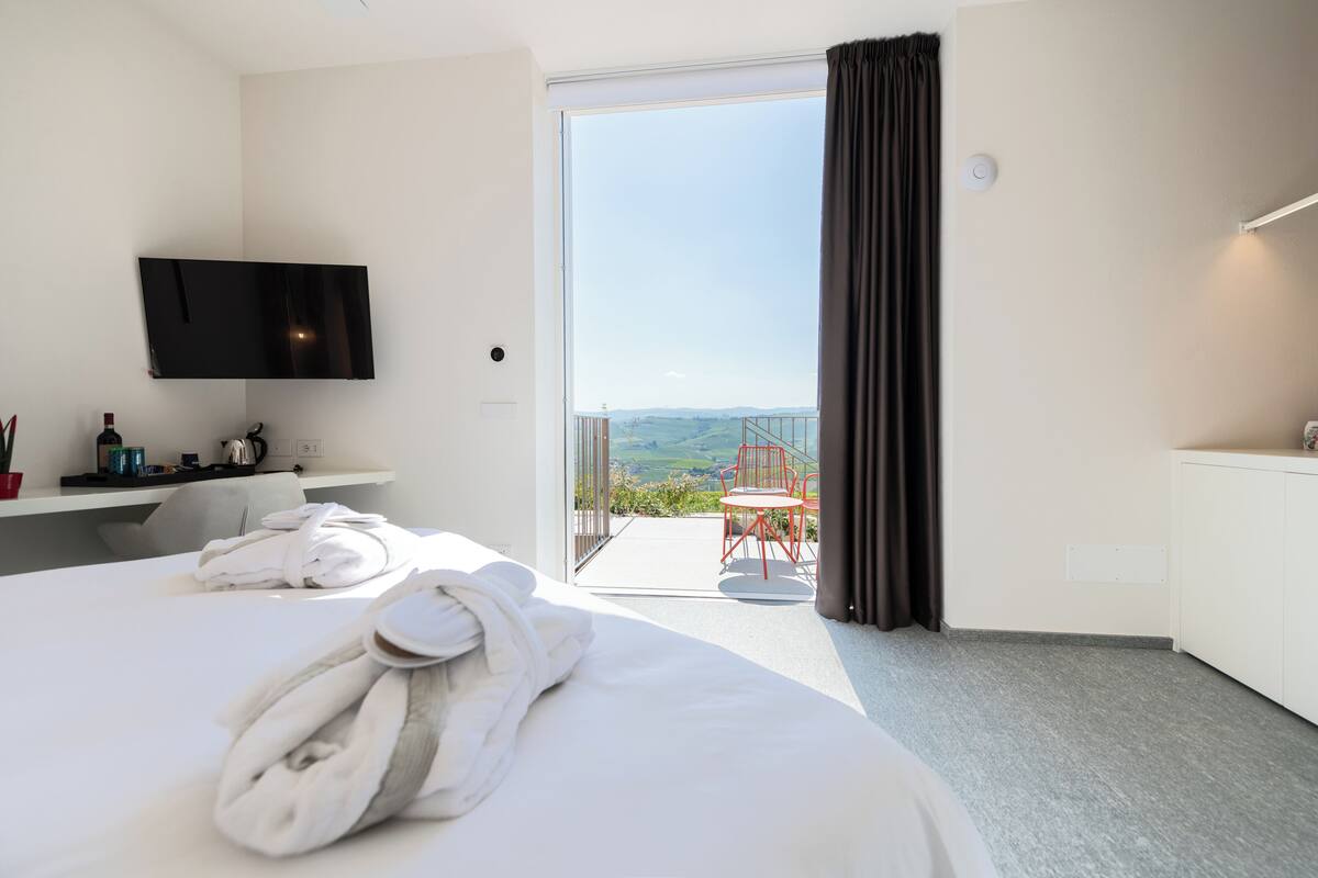Habitación de hotel con cama, televisor montado en la pared, una mesita con hervidor, una silla roja y un balcón con vistas al campo.