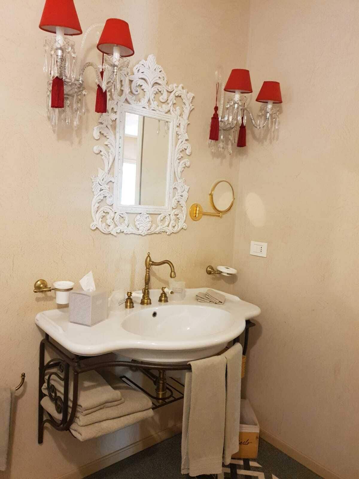 Une salle de bain avec un lavabo blanc, des robinets dorés, un miroir doté d’un cadre orné et des abat-jours rouges.