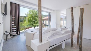 A modern bedroom with a large bed, a desk, and a chair. There is a window with a view of a tree and buildings. - Palas Cerequio BaroloCruResort (Barolo)