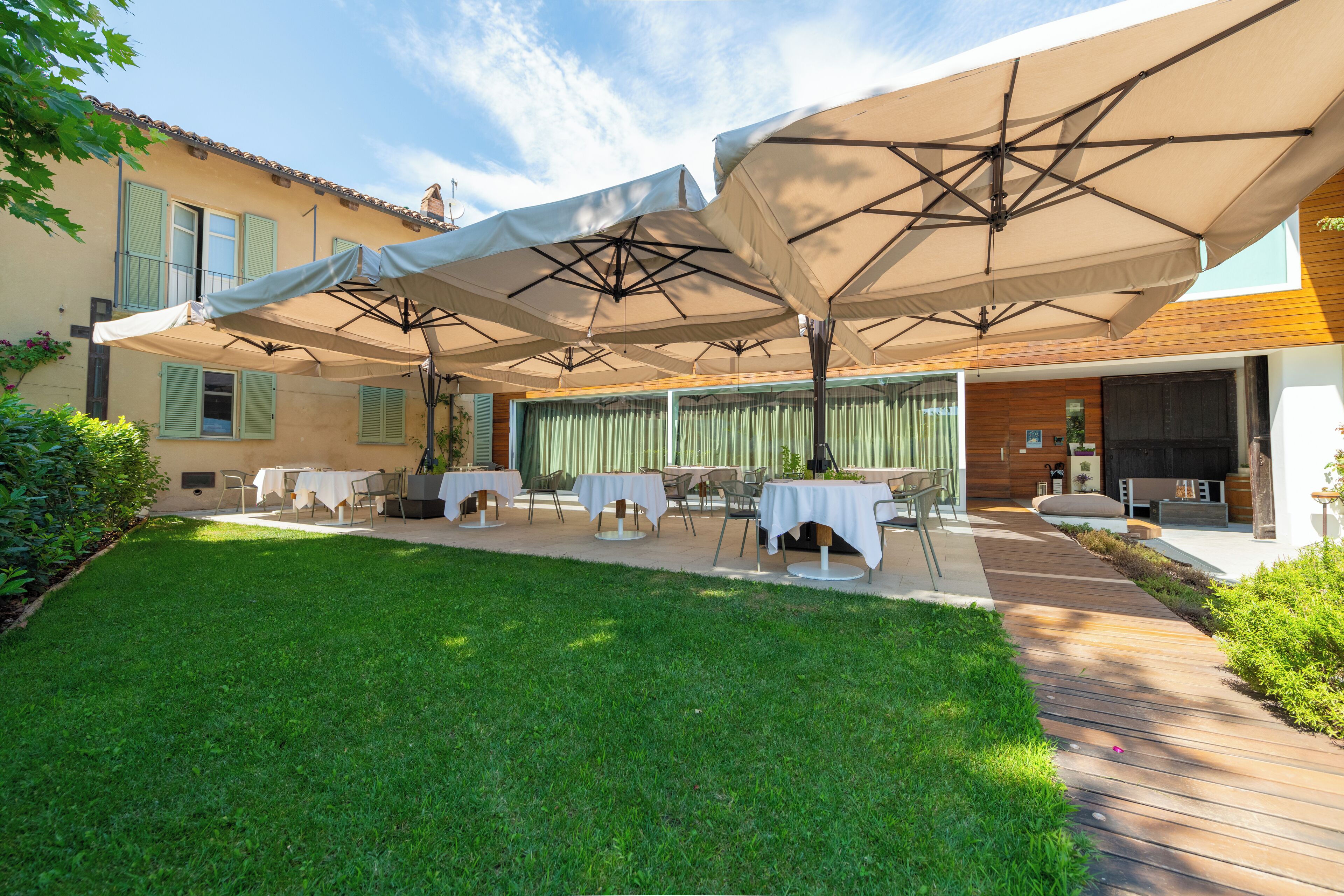 An outdoor patio with tables and umbrellas, surrounded by a well-maintained lawn and a building with a tiled roof.