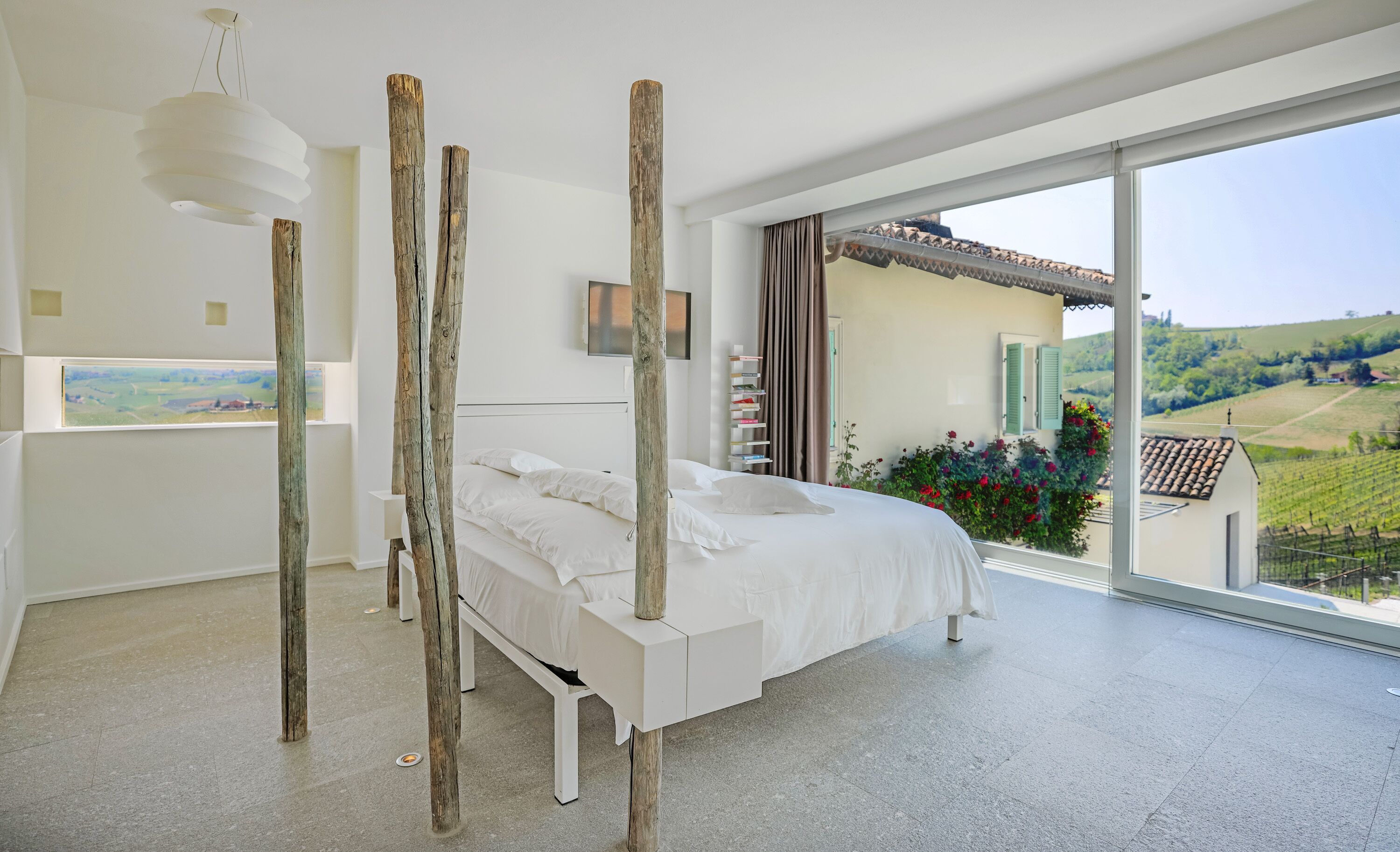 A modern bedroom with a large bed, wooden pillars, a view of the countryside, and a balcony with plants.
