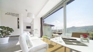 A modern hotel room with a large window, a white sofa, a dining table with wine glasses, and a potted plant. - Palas Cerequio BaroloCruResort (Barolo)