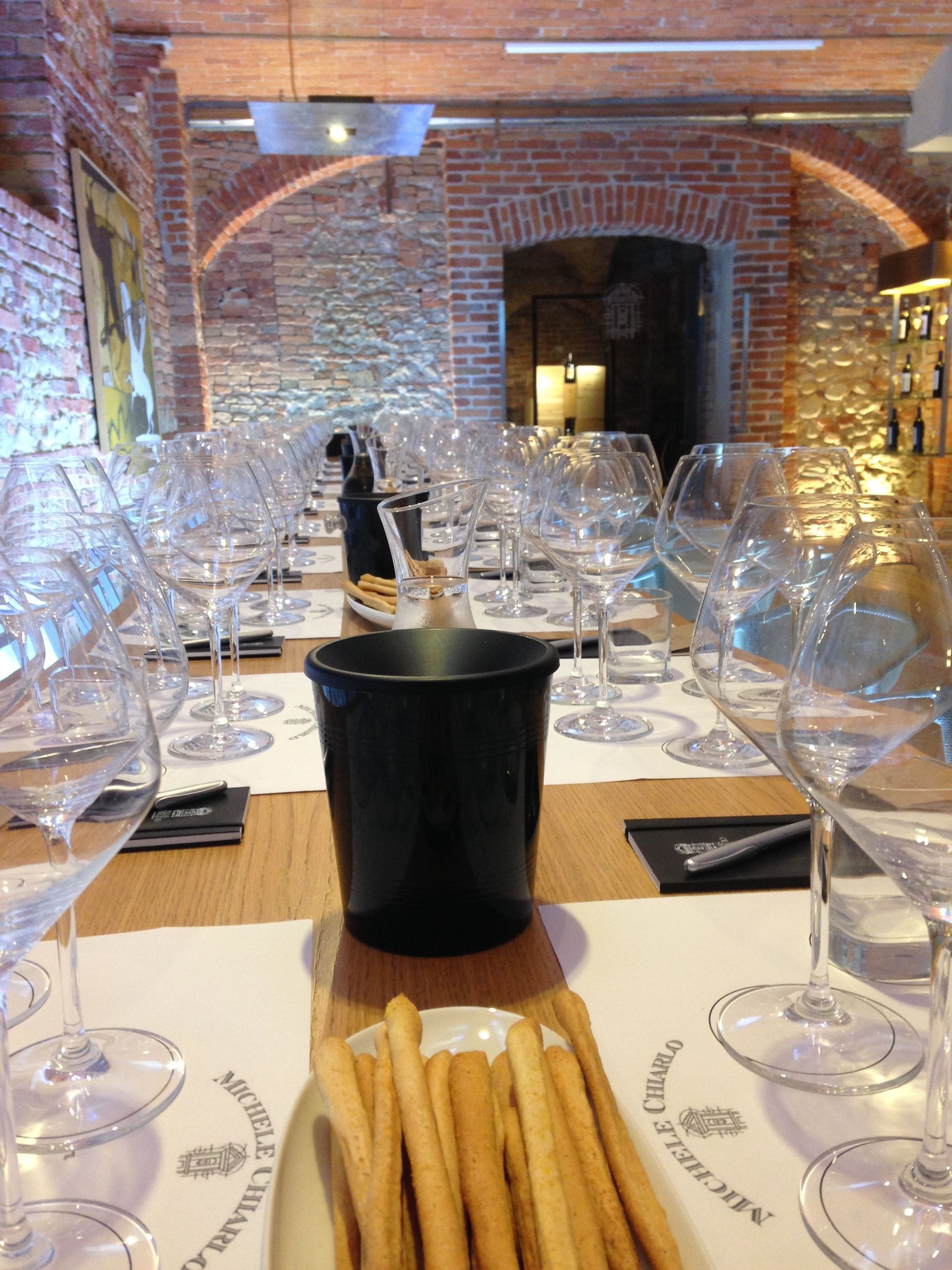 A restaurant setting with a long table set for a wine tasting event, featuring numerous wine glasses and a black bucket in the center.