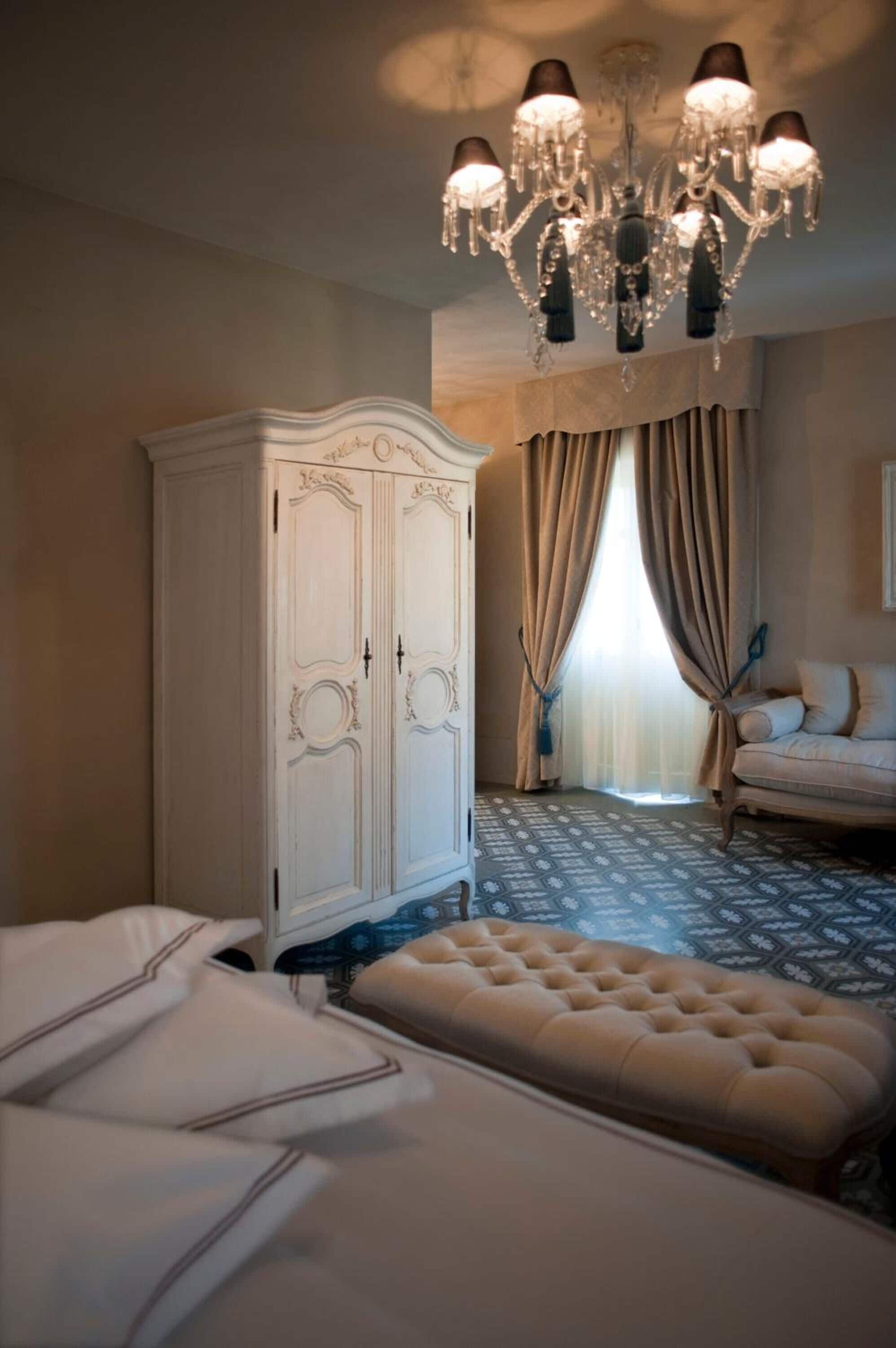 Quarto com um guarda-roupa branco grande, um banquinho estofado, um candelabro e um tapete estampado.