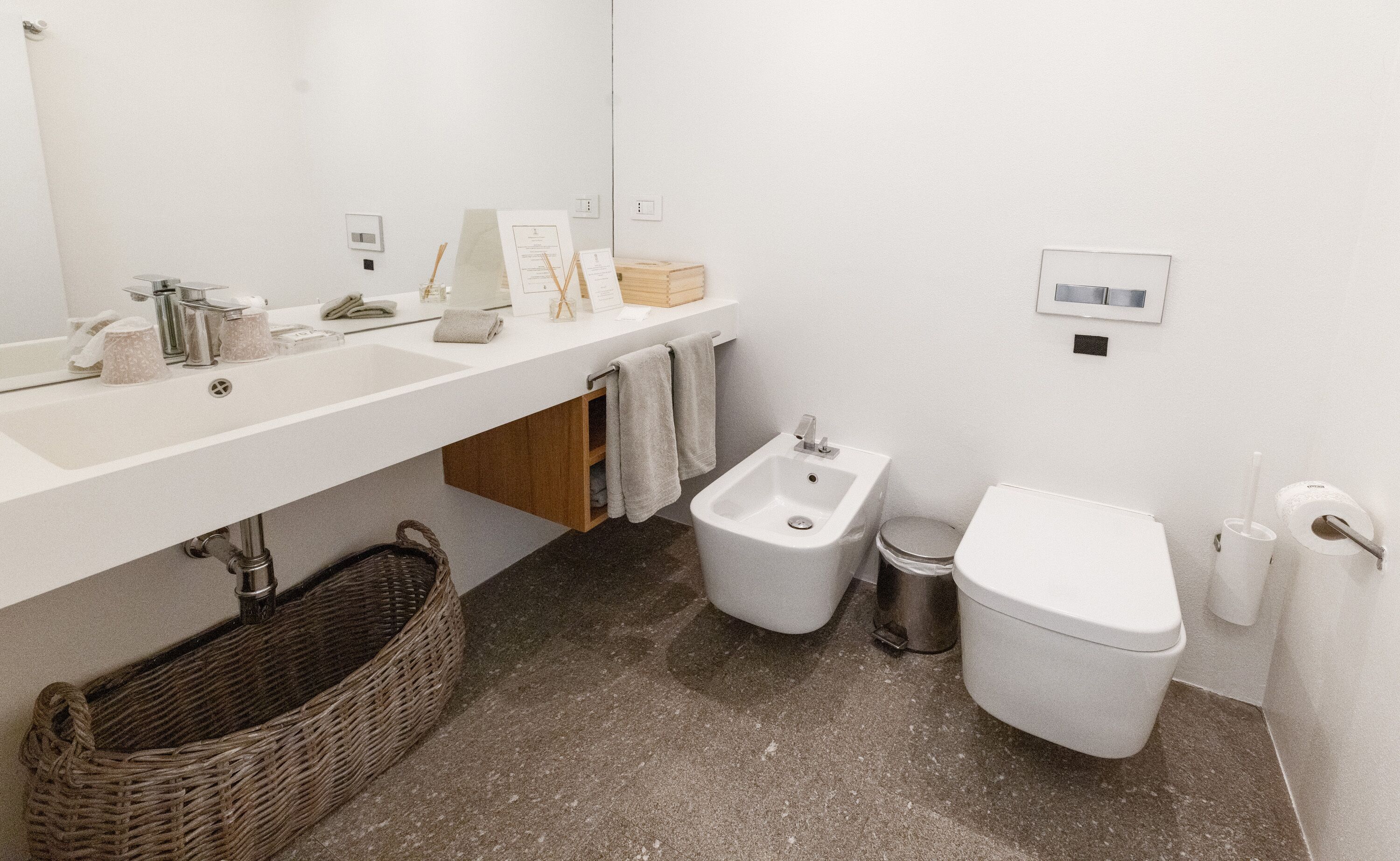 A modern bathroom with a white sink, bidet, and toilet. There is a wicker basket under the sink and a trash can next to the toilet.