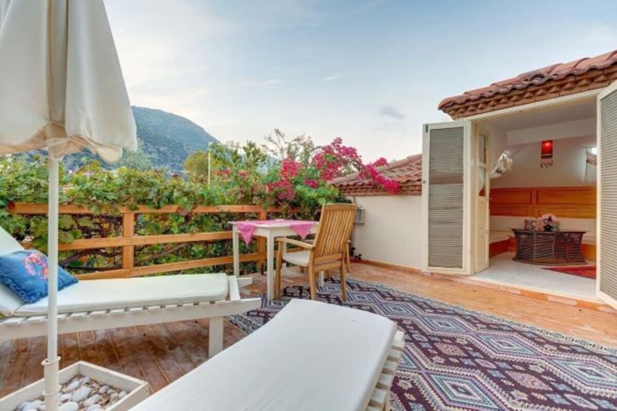 A balcony with a white sofa, a table, and a chair. There is a wooden fence and a view of mountains.