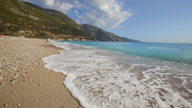 Una playa con piedras y olas, montañas al fondo y un cielo despejado.
