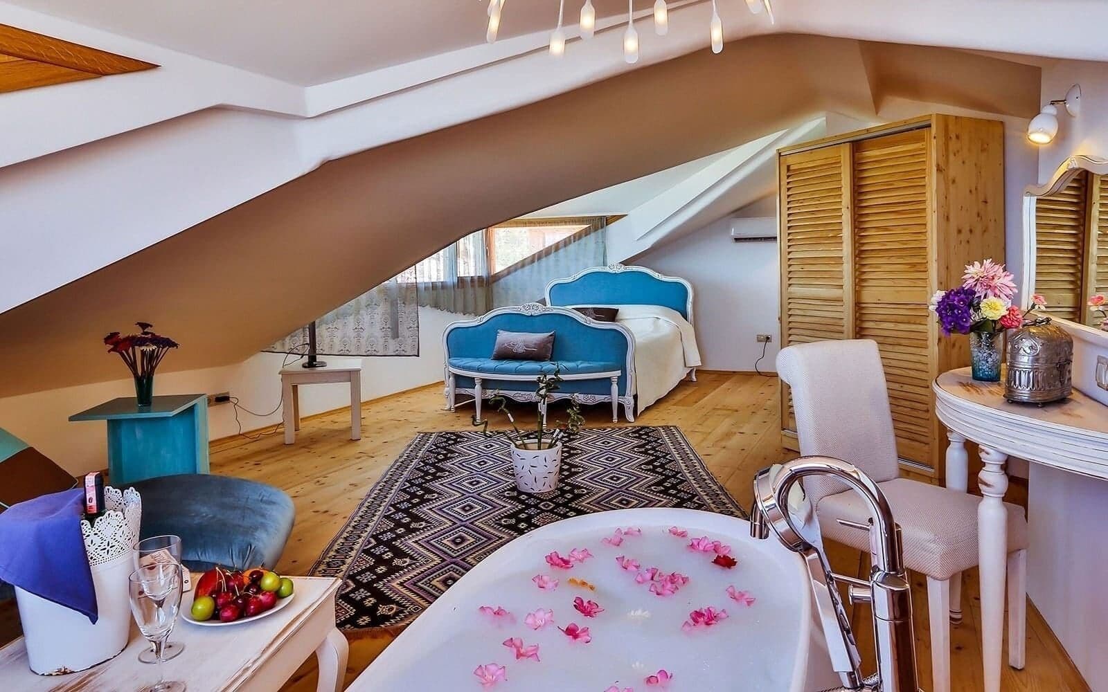 A cozy loft bedroom with a sloped ceiling, a large window, a blue sofa, a white chair, a bathtub with rose petals, and a small table with a vase of flowers.
