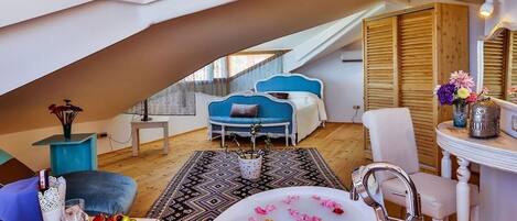 A cozy loft bedroom with a sloped ceiling, a large window, a blue sofa, a white chair, a bathtub with rose petals, and a small table with a vase of flowers.