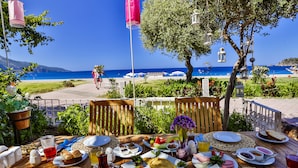 Una mesa dispuesta con alimentos para el desayuno, como huevos, pan y fruta, con vista a una playa con sombrillas y un cielo azul despejado.