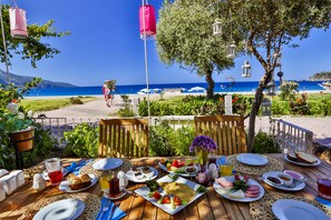 Ein Tisch reichhaltig mit Frühstück gereicht, darunter Eier, Brot und Obst, mit Blick auf einen Strand mit Sonnenschirmen und einem klaren, blauen Himmel.