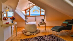 Un dormitorio acogedor en el ático con una bañera grande, un espejo, una mesita con un florero y vistas al mar a través de una ventana.