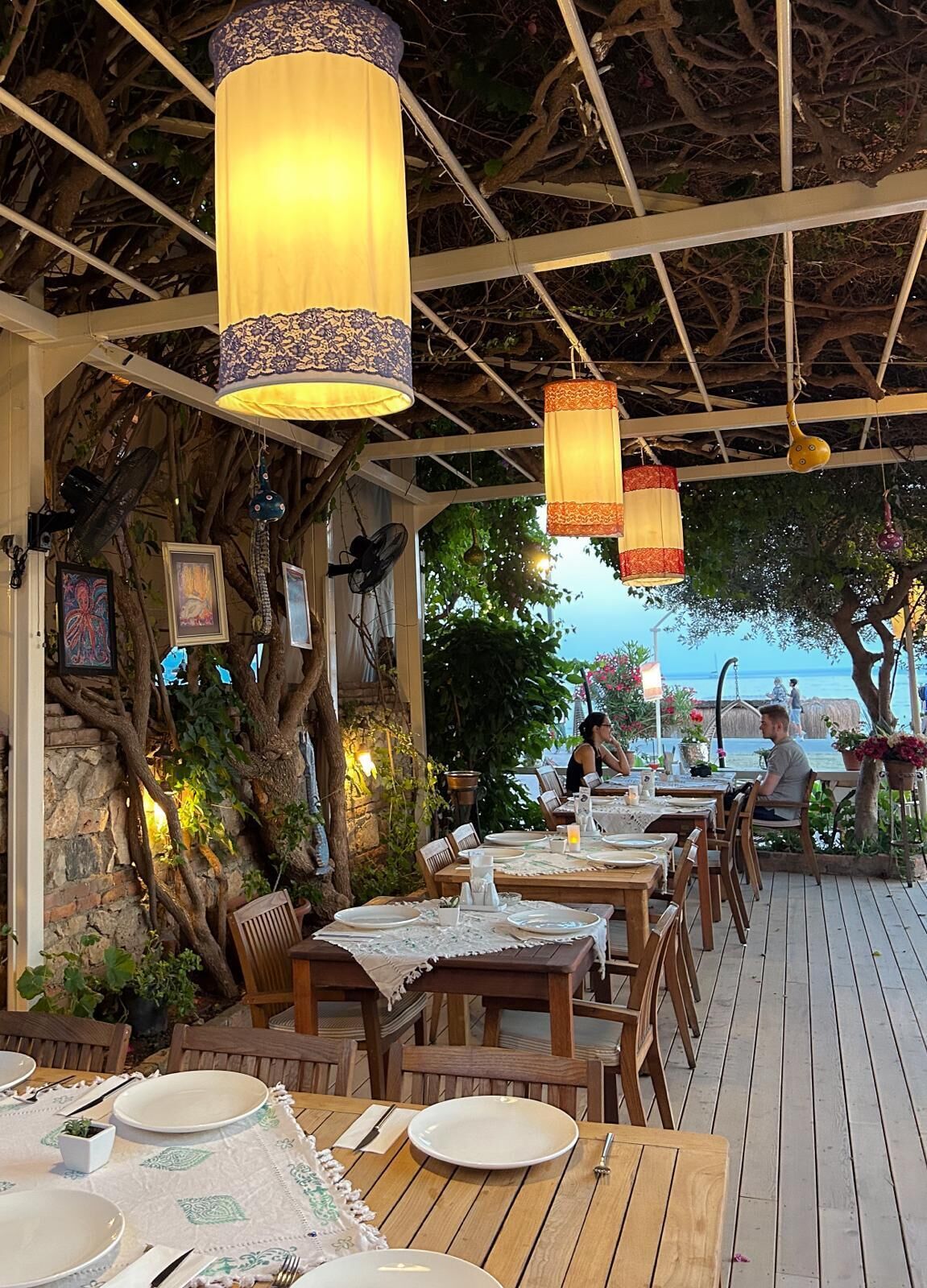 An outdoor dining area with wooden tables and chairs, hanging lanterns, and a view of the sea.