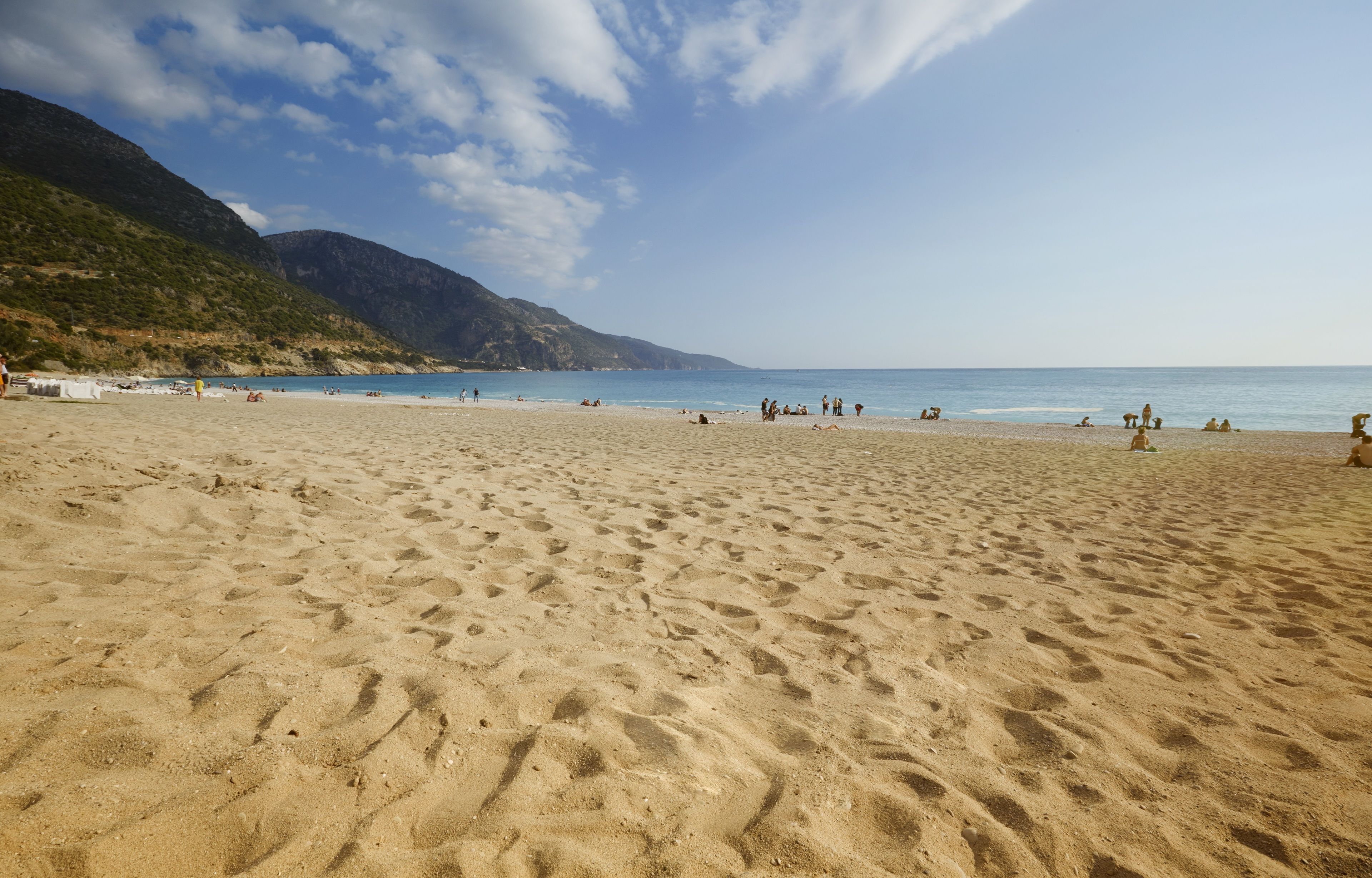 Een breed zandstrand met mensen die zonnen en zwemmen, omringd door bergen en een heldere lucht.