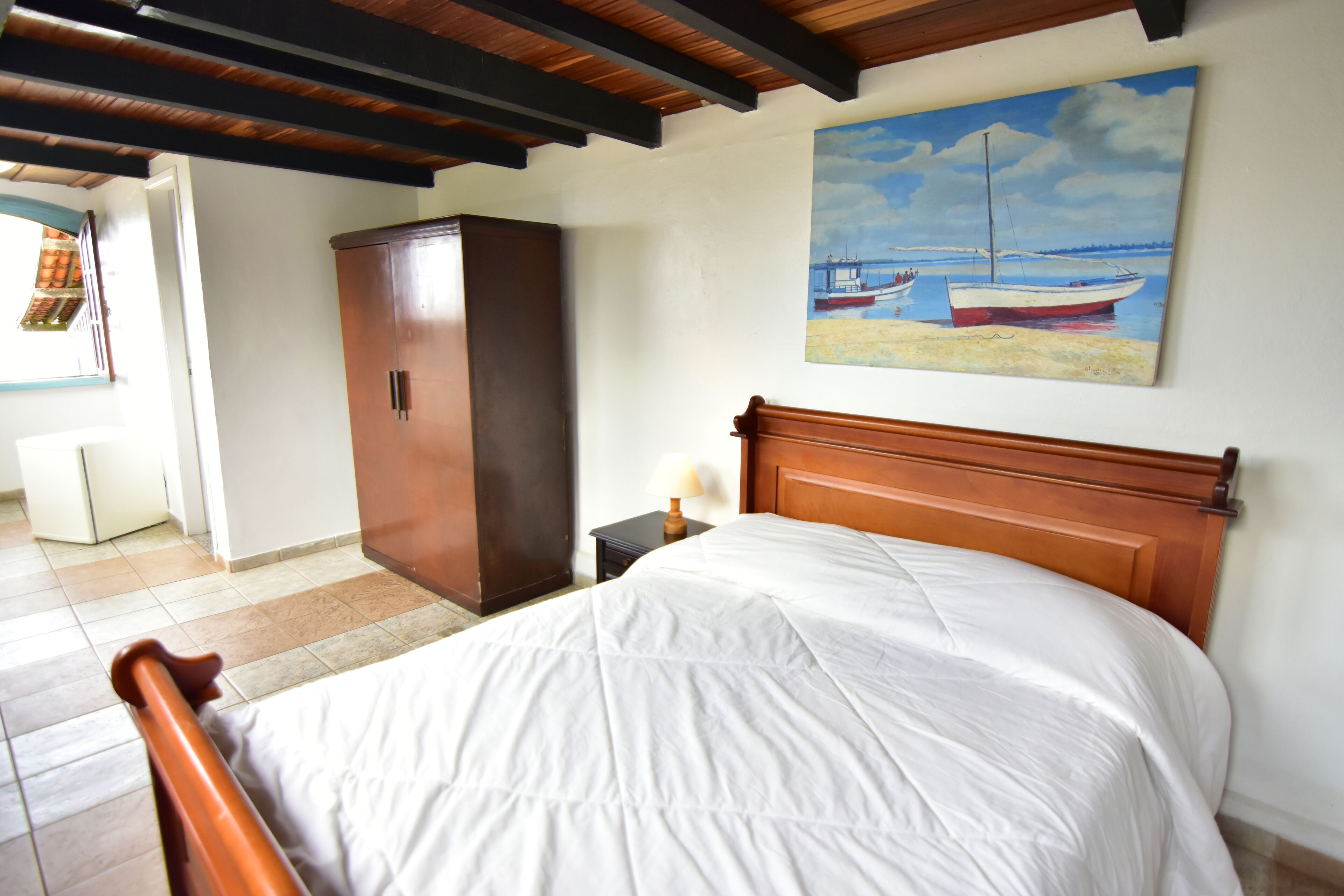 A bedroom with a wooden bed, a nightstand with a lamp, a wardrobe, and a painting of boats on the wall.