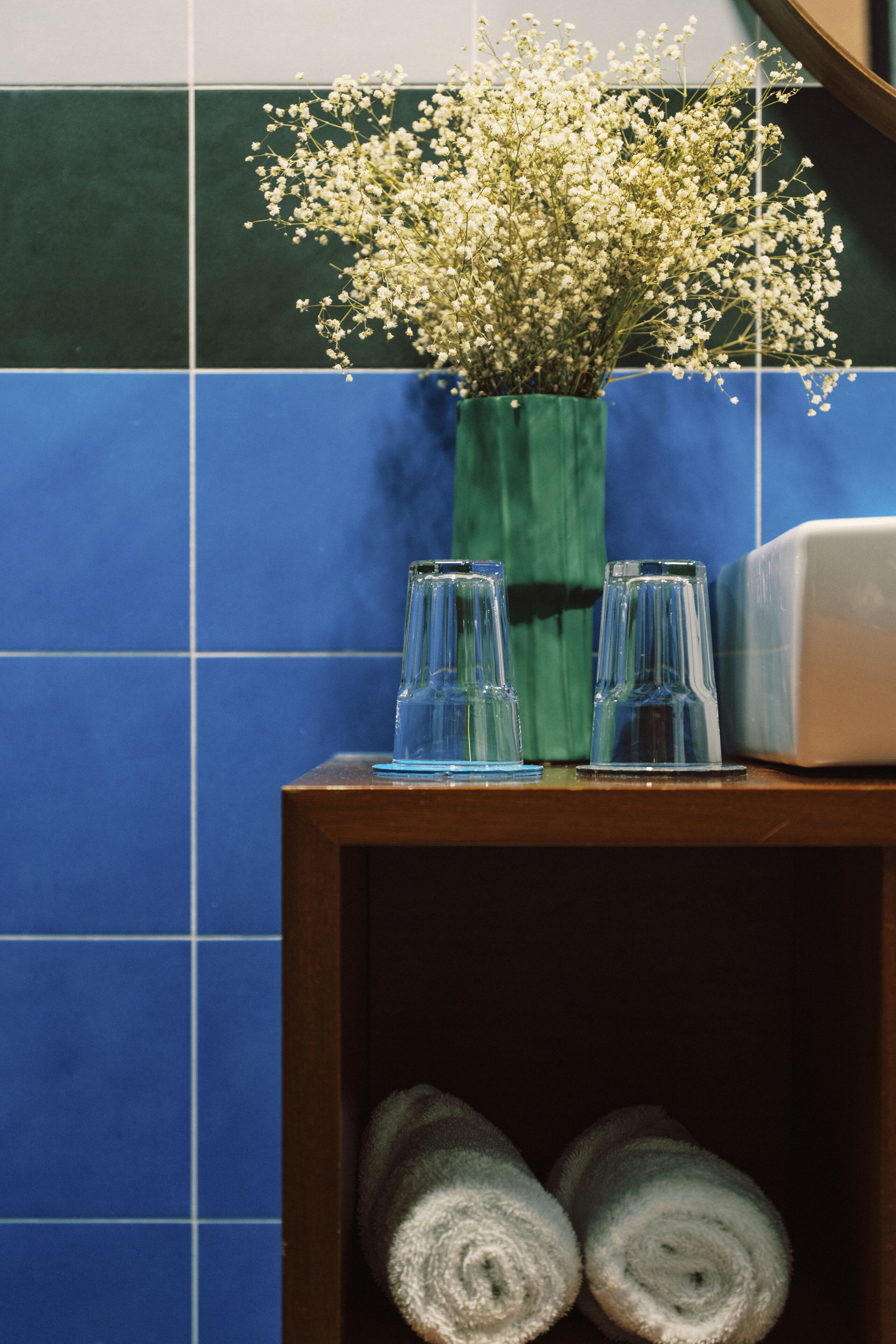 A bathroom with a blue and green tiled wall, a vase with white flowers, two clear glasses, and a towel rack with rolled towels.
