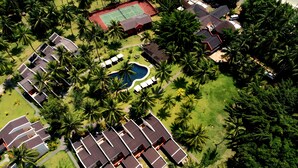 Vista aérea de um resort com várias construções no estilo de bangalôs, uma quadra de tênis e uma piscina cercada por palmeiras.