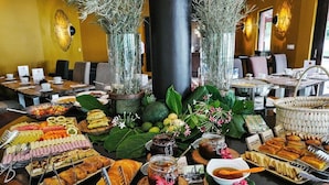 Uma mesa de buffet com diversos pratos e bebidas, cercada por plantas decorativas e um pilar central.