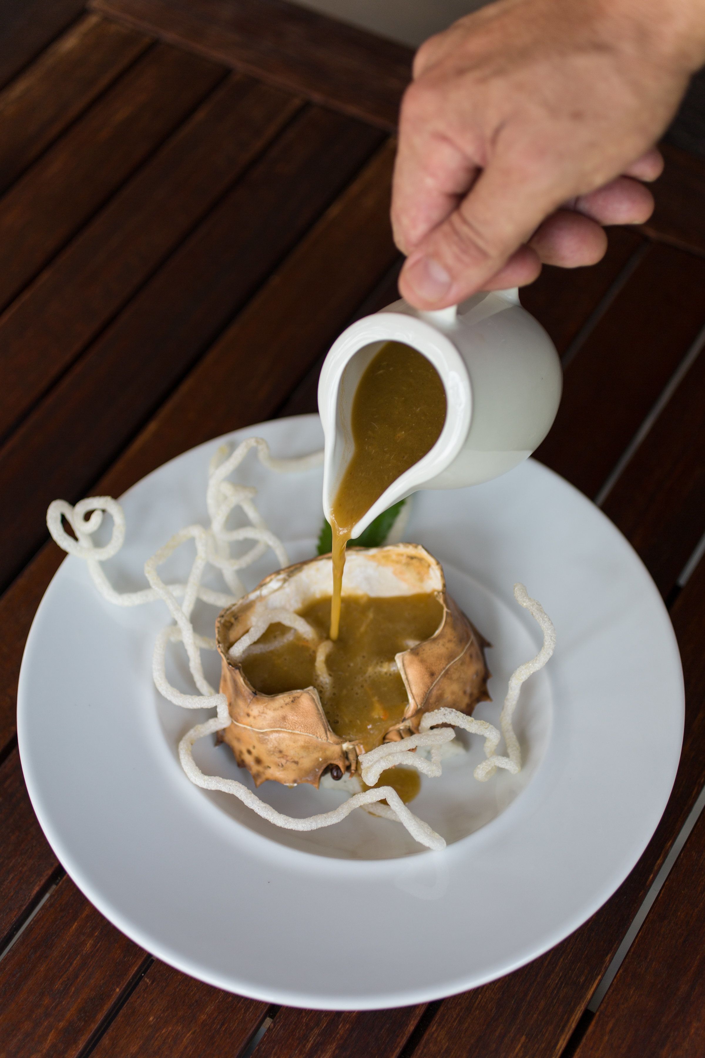 A hand pouring sauce into a dish with noodles and a piece of meat on a white plate.
