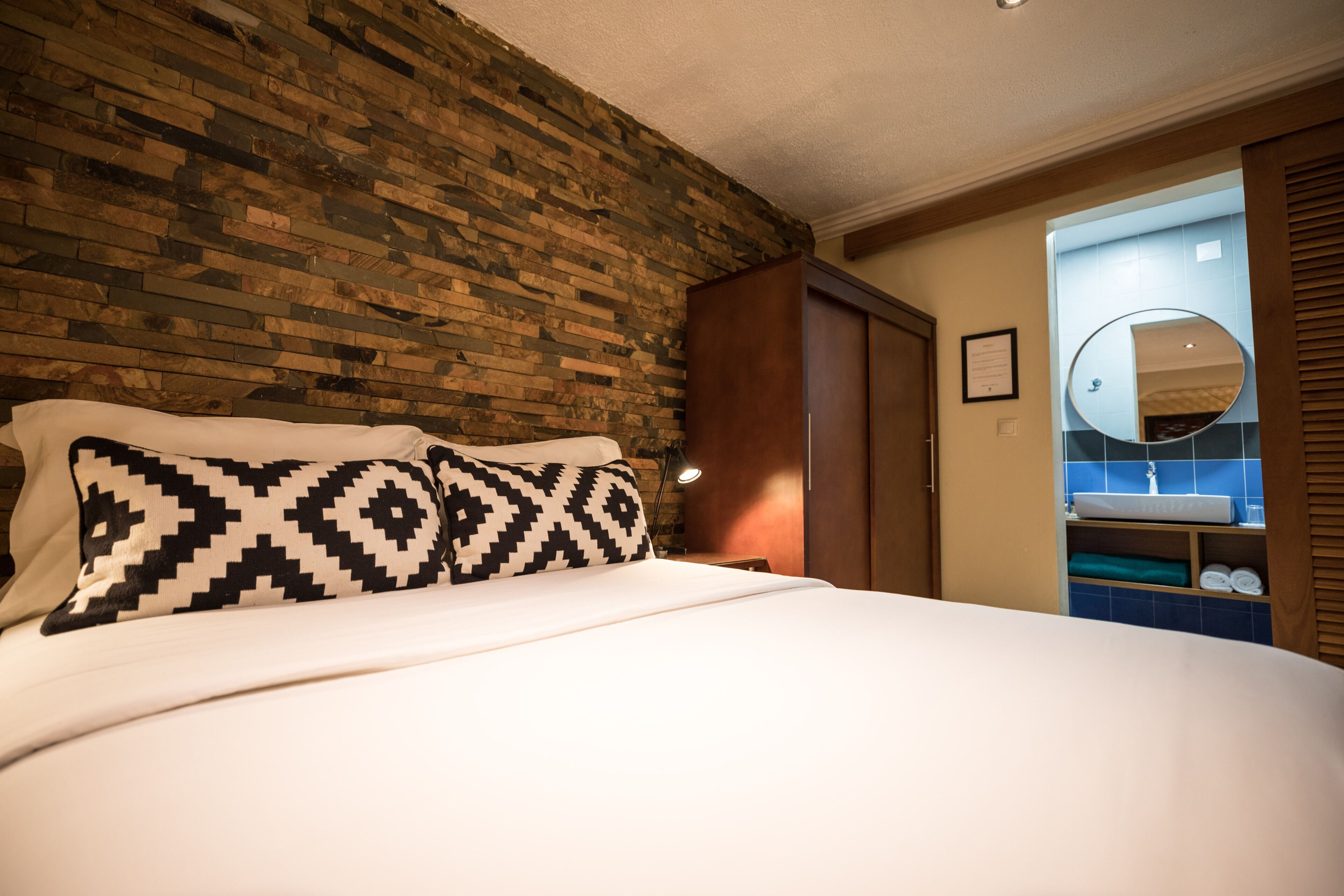 Habitación de hotel con pared de ladrillo, cama con almohadas estampadas, armario de madera y baño visible a través de una puerta abierta.