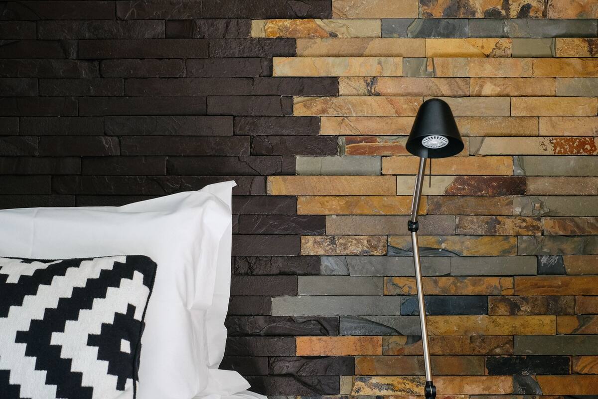 Ein Bett mit einem weißen Kissen und einem Kissen in Schwarz-Weiß-Muster, eine schwarze Stehlampe und eine Wand aus einer Mischung aus dunklen und hellen Steinen.