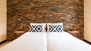 Cama de casal com roupa de cama branca e almofadas estampadas em preto e branco, apoiada contra uma parede de tijolos.
