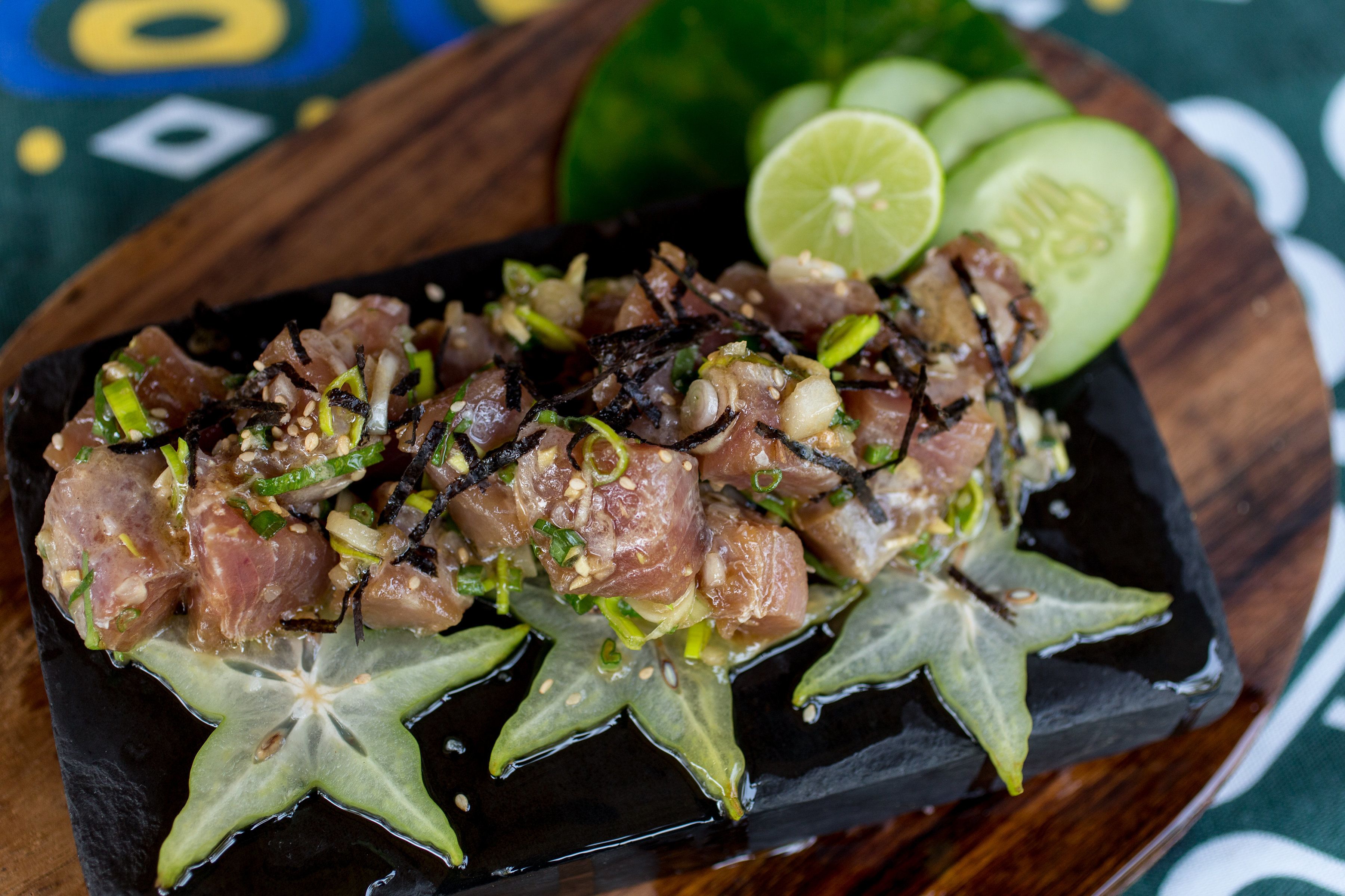 A dish of raw fish topped with green onions and sesame seeds, served on a black plate with star-shaped leaves and lime wedges.