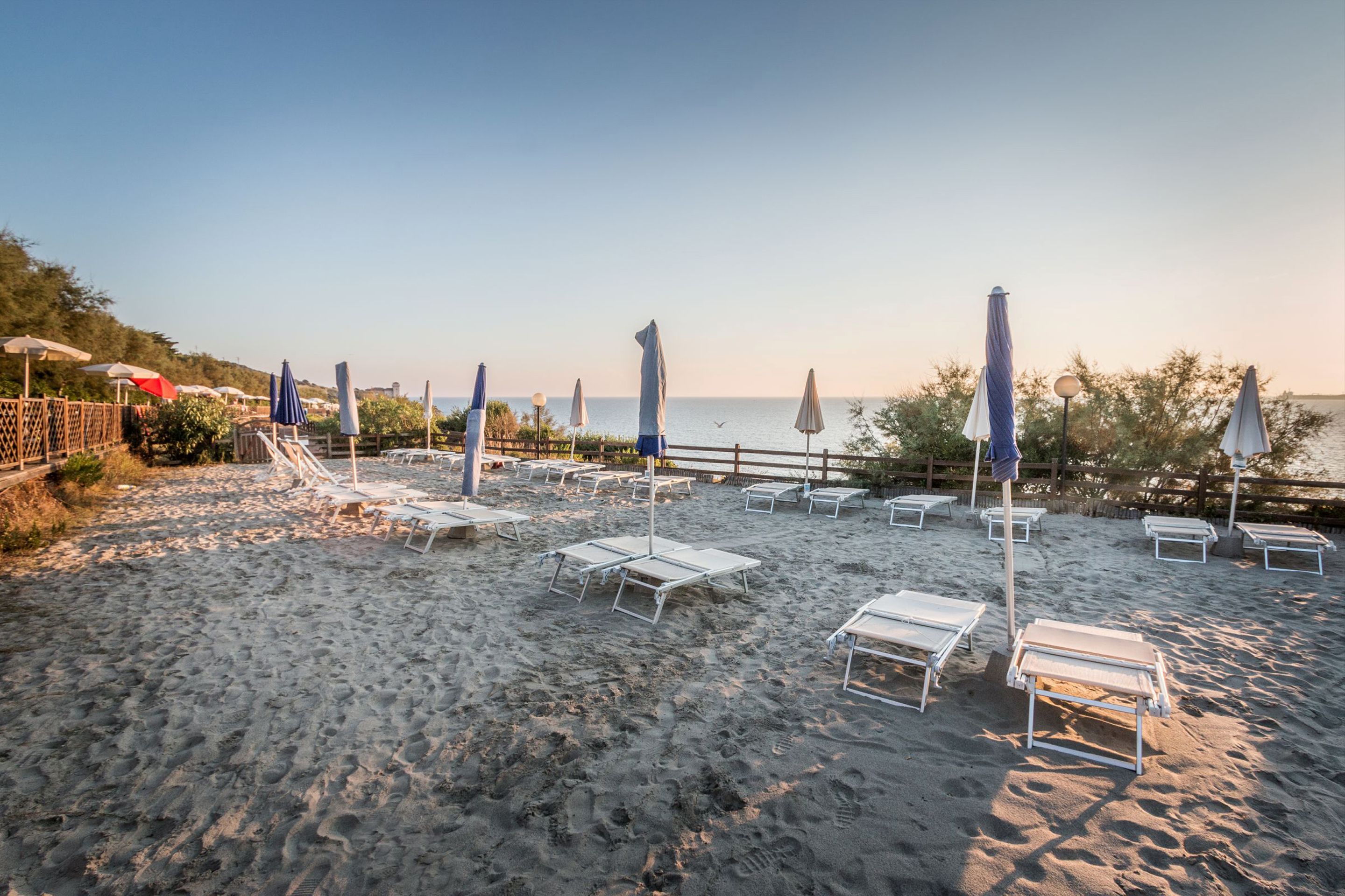 A beach with sun loungers and umbrellas, a wooden fence, and a clear sky.
