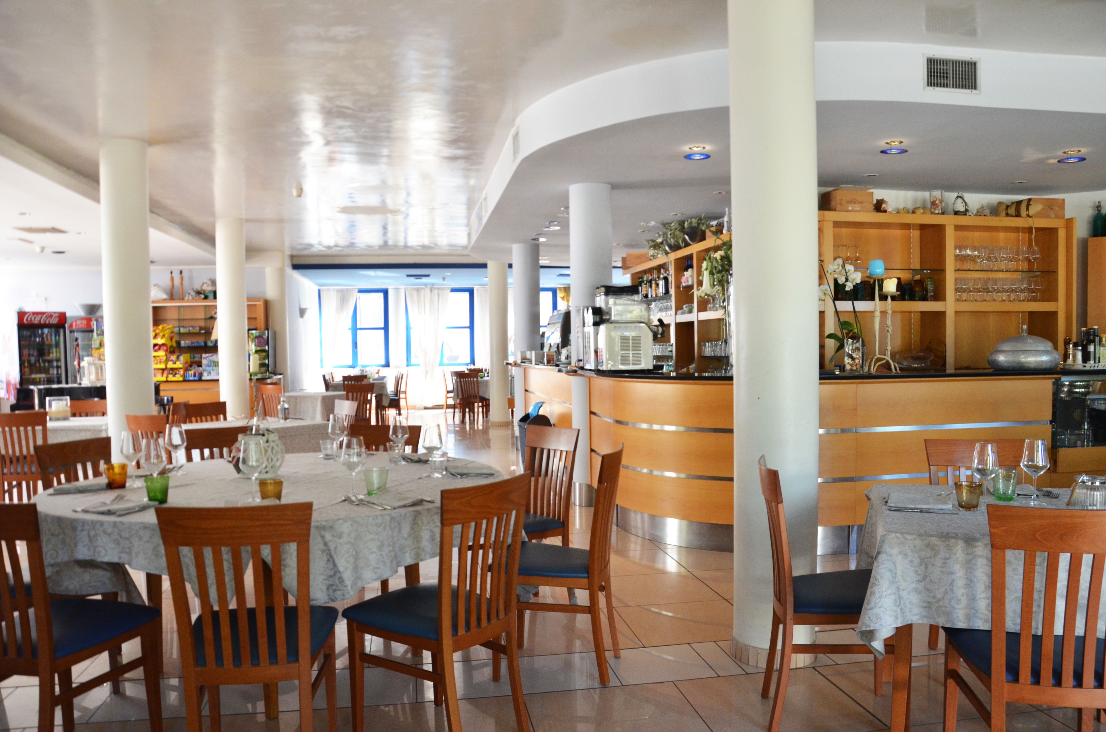Un restaurant spacieux avec des tables rondes et des chaises en bois, un bar avec des étagères remplies de bouteilles, et une vue sur l’extérieur grâce à de grandes fenêtres.