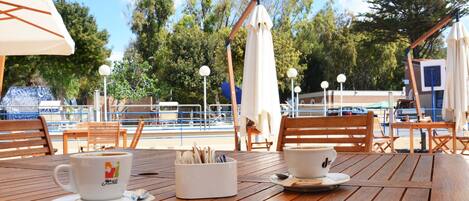 A wooden table with two white cups and a small white container, likely for napkins, in a sunny outdoor setting with umbrellas and a pool in the background.