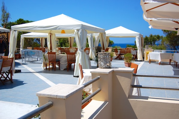 Una terraza al aire libre con toldos blancos, mesas y sillas de madera, con vistas al mar.
