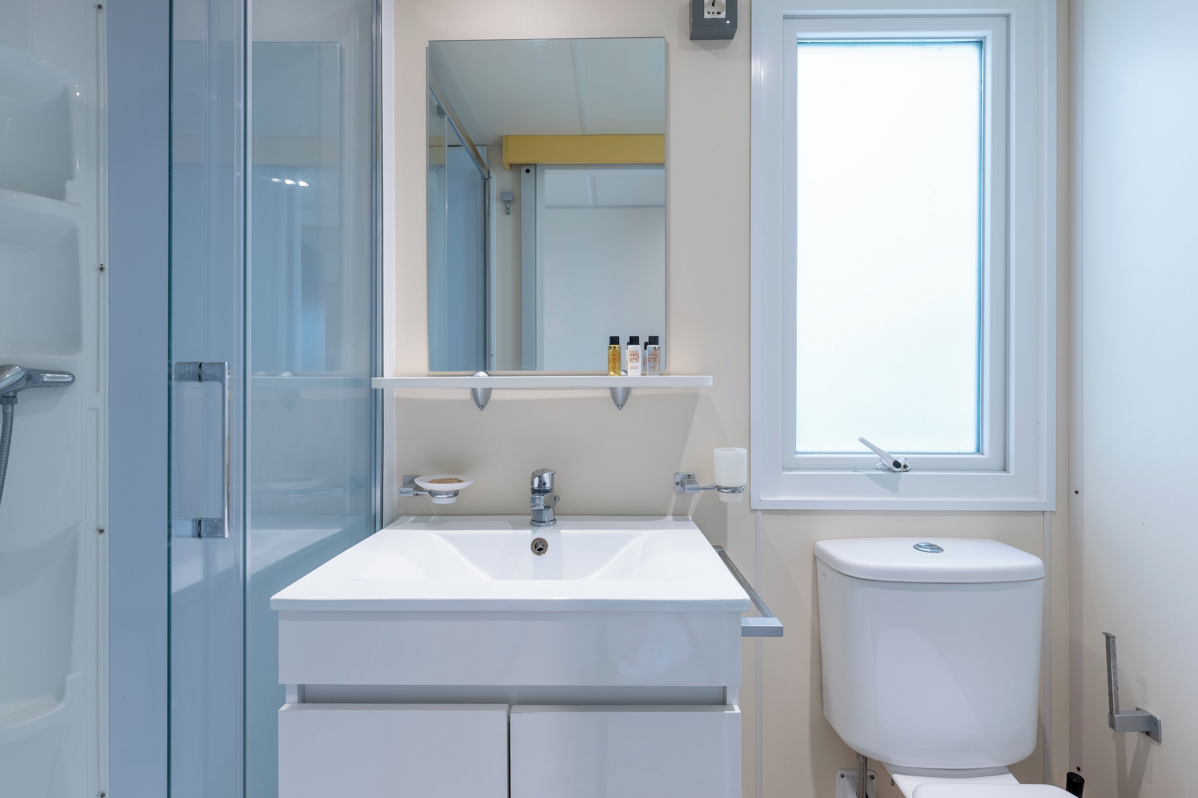 A modern bathroom with a glass shower enclosure, a white sink with a mirror above it, and a toilet with a window above.