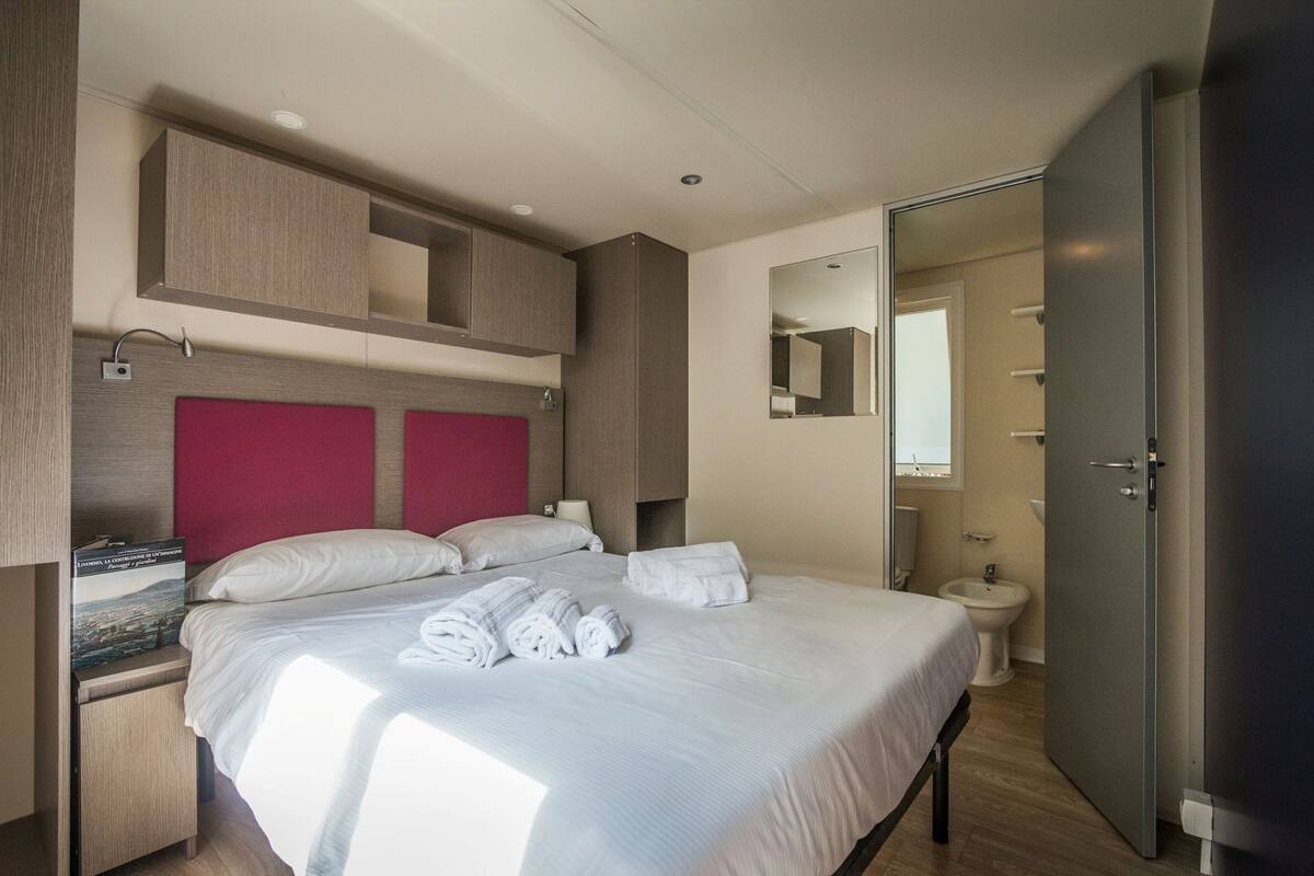A hotel room with a large bed, a red headboard, a wardrobe, a bathroom with a mirror, and a toilet.