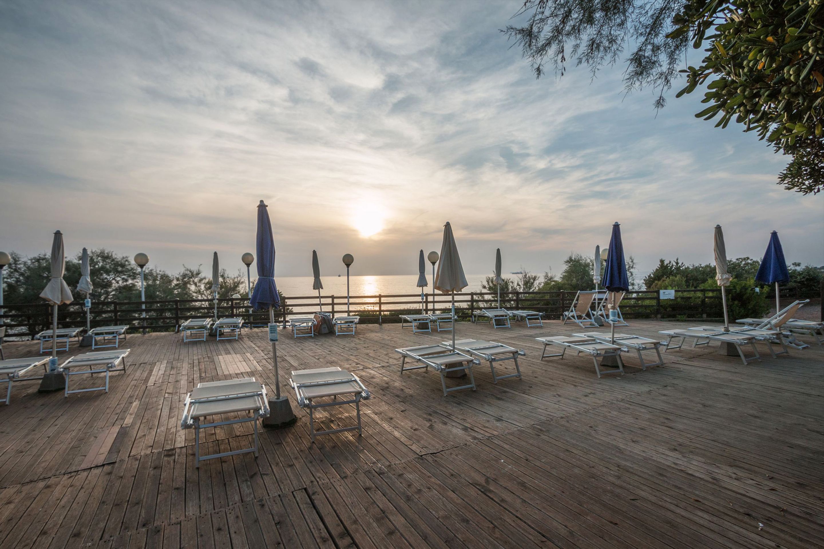 A wooden deck with lounge chairs and umbrellas, overlooking a body of water with a sunset in the background.