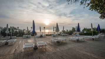 A wooden deck with lounge chairs and umbrellas, overlooking a body of water with a sunset in the background.