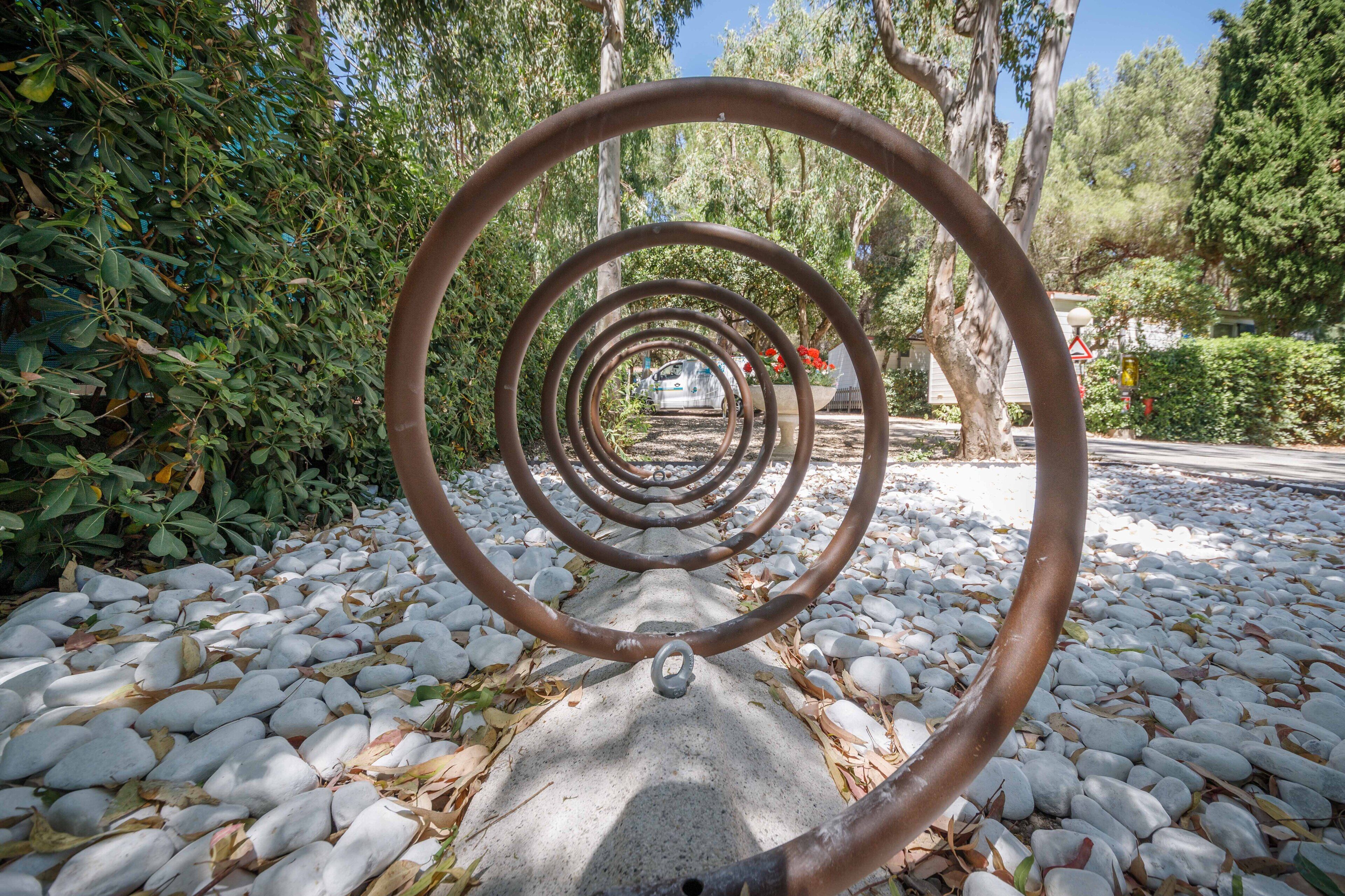 A spiral metal sculpture in a park with trees and white pebbles on the ground.