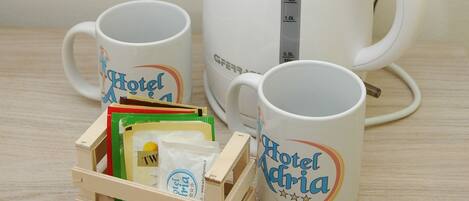 Una tetera eléctrica blanca, dos tazas blancas con la inscripción 'Hotel Maria' y una caja de madera con varios objetos, incluyendo una tarjeta verde y un paquete blanco.