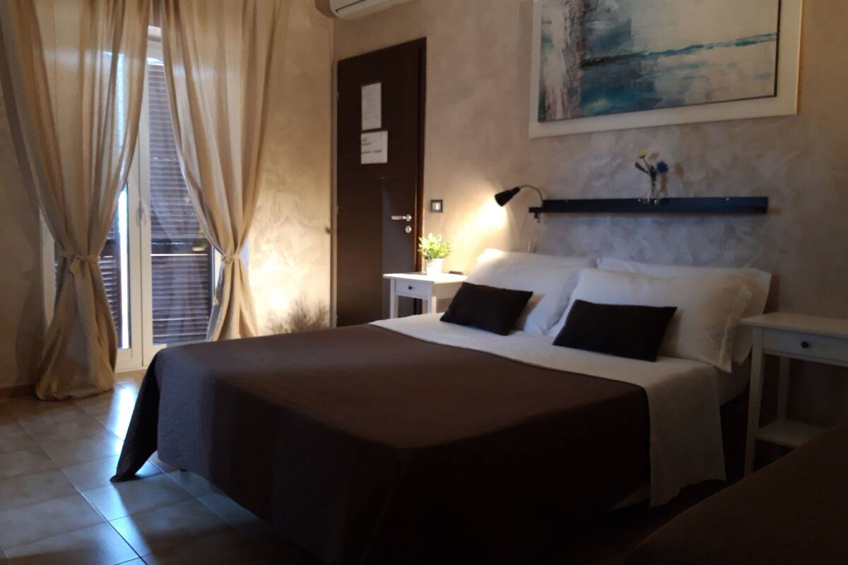 Een hotelkamer met een groot bed, een nachtkastje, een wandairco, een schilderij en een raam met gordijnen.