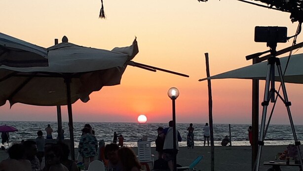 A beach scene with people sitting at tables under umbrellas, a sunset in the background, and a camera on a tripod. - B&B Giorgia Airport (Fiumicino)
